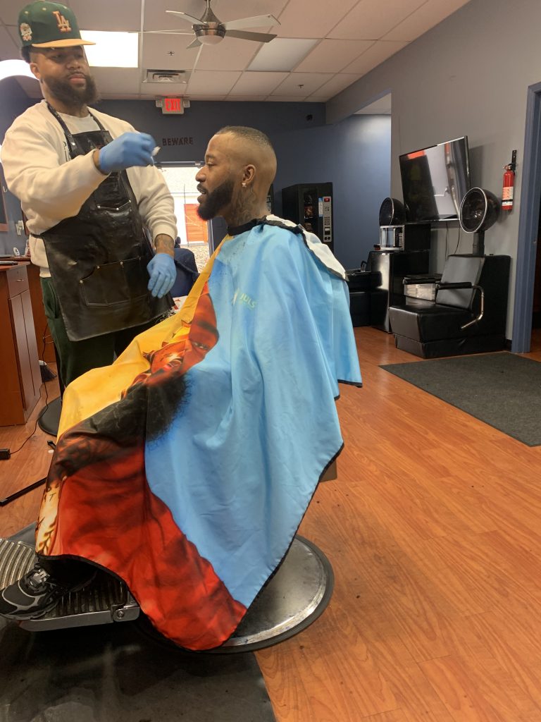 A man sitting on the barber chair getting his haircut. The barber is standing in front of him.