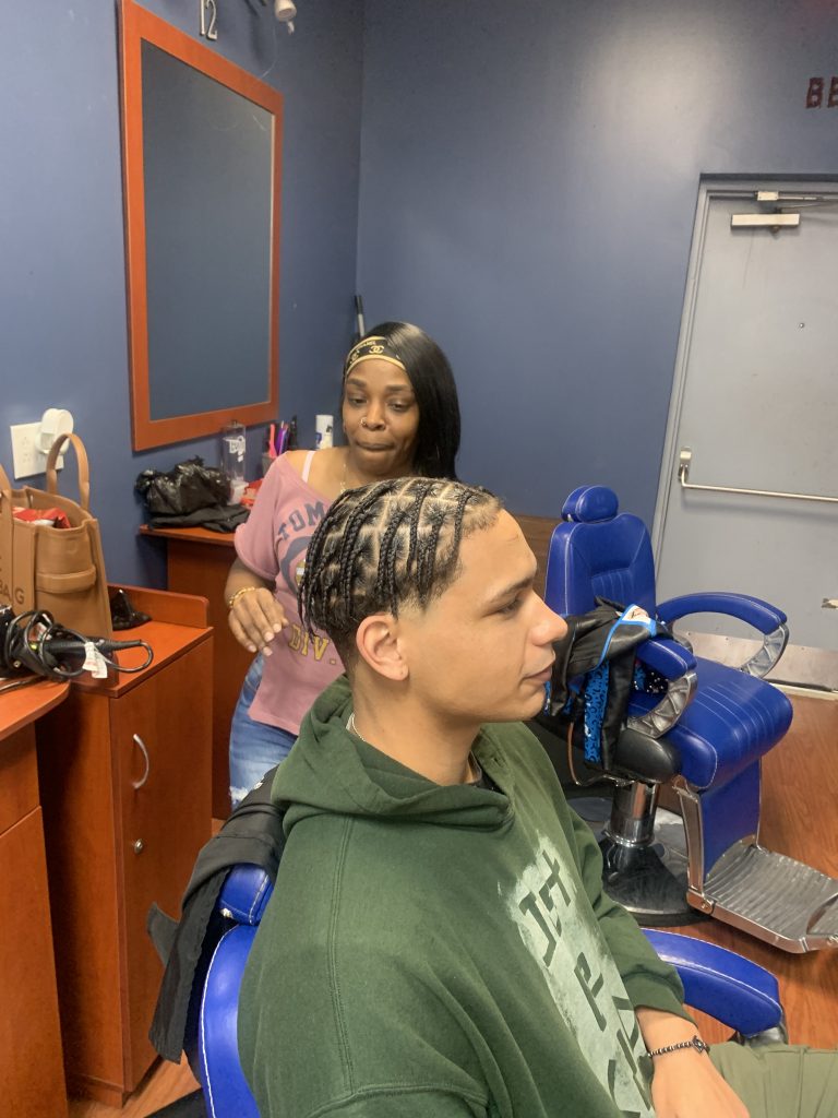 A male customer sitting on a barber chair and the hairstylist standing behind him looking at the braided hair.
