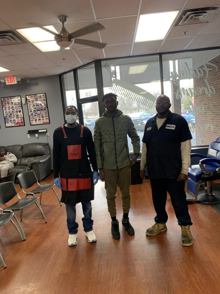 Three males standing next to one another. The barber and two clients.