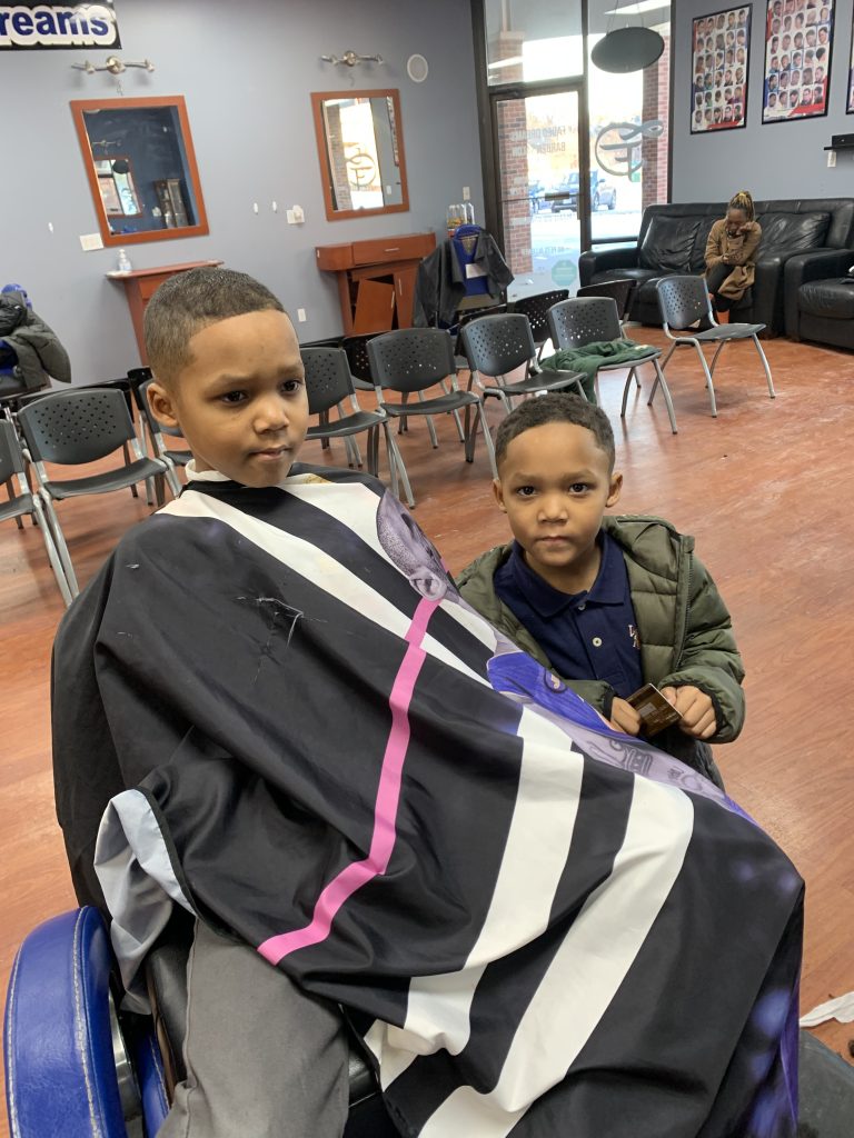 Two boys after their haircut. One sitting on the barber chair and one standing next to him.