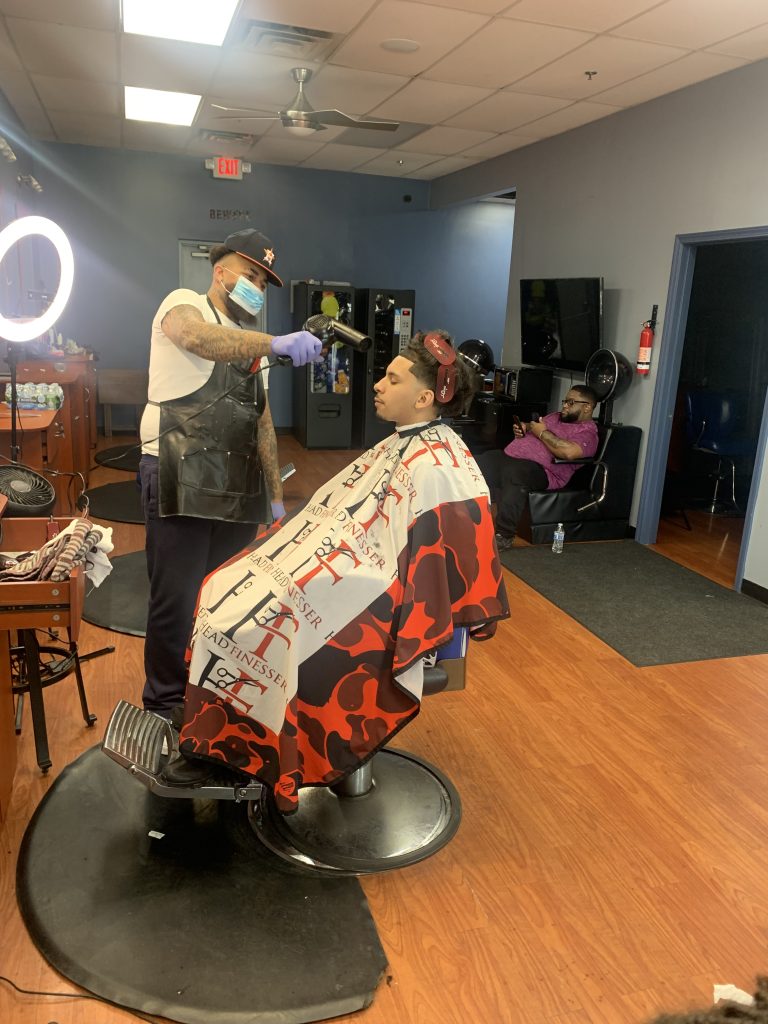 Babber using a blowdryer on a male client that is sitting on a barber chair.
